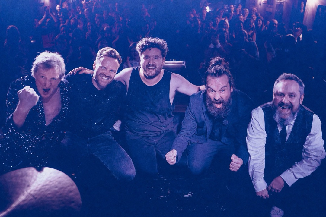 Die komplette Band steht mit erhobenen Armen im Bühnenlicht.